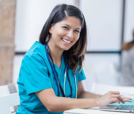 A female Filipino nurse is sitting down at her laptop to study. She is wearing scrubs and a stethoscope. She is smiling at the camera. Image credit: SDI Productions / E+ / Getty Images