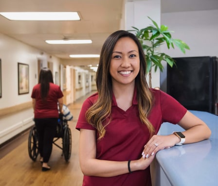 Asian-American female medical assistant smiles while standing in front of a hospital reception desk.