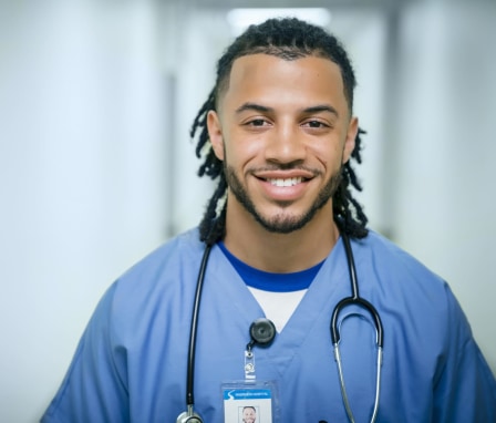 A young mixed race nurse is standing in a hospital hallway. He is wearing light blue scrubs and is looking at the camera and smiling.