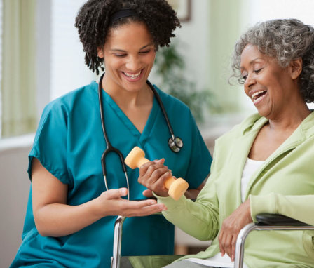 A mid-adult African-American home nurse is helping her elderly female patient in a wheelchair lift a hand weight. They are sitting together in the patient's home living room.