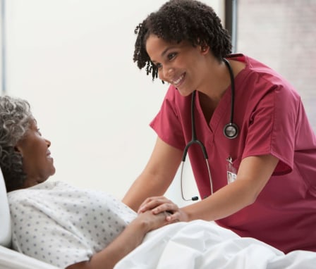 A young Black female nurse is checking in on one of her elderly patients during her hospital rounds. She has short, curly black hair and is wearing pinkish-red colored scrubs. The patient is an elderly black female wearing a hospital gown. The nurse is standing beside the patient's bedside, smiling and chatting with her.