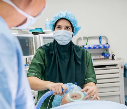 A female nurse anesthetist places an oxygen mask on a senior patient while sedating him in preparation for a surgical procedure in a hospital operating room. The young Malaysian nurse is wearing a dark green hijab over her green scrubs, and has a mask and medical cap on. A heart monitor and medical equipment are in the background. The nurse is looking towards the doctor in the foreground while awaiting his instructions.