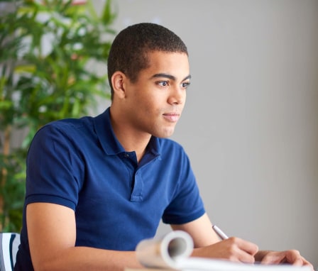 A young man sitting and taking a practice exam at his desk. He is writing down his test answers on a notepad while looking at his computer.