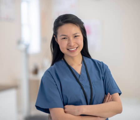 A young Asian-American female wearing blue medical scrubs smiles at the camera.