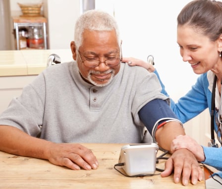 A mid-adult Caucasian female nurse is at an elderly Black patient's home taking his blood pressure. They are sitting at the patient's dining room table and smiling.