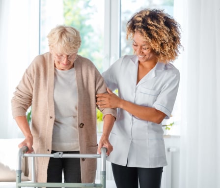 A cheerful African-American nurse is helping a senior female patient to use a walking frame. The nurse has her arms around the patient's left shoulder and back to offer support. They are both inside the patient's home living room.