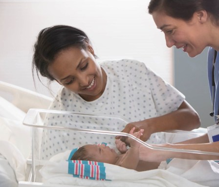 A mid-adult female nurse has just carted a newborn child to its mother's hospital bedside. The mother is a mid-adult Black woman wearing a hospital gown. She is smiling and leaning over her bedside to greet her newborn child.