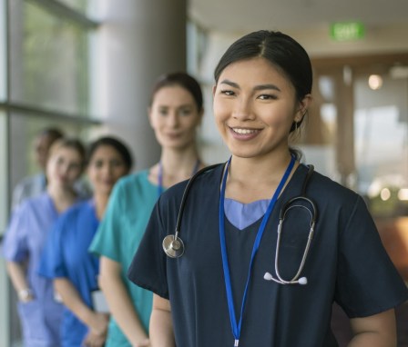 A female doctor of Asian descent poses with her multi-ethnic team of doctors and nurses and smiles directly at the camera while standing in a hospital corridor.