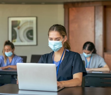 Group of nursing students wearing protective face masks back at school take notes while attending class lecture. Each individual is seated at their own desk due to Covid-19 social distancing regulations.