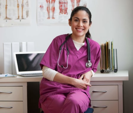 A female Hispanic nurse sitting at her desk in a clinical office. She is facing towards the camera and smiling. Her laptop and case files are on the desk behind her.