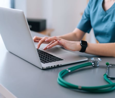 Female nurse in scrubs sitting at her home desk is typing on her laptop keyboard