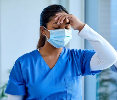 Young Asian-American female nurse with her eyes closed rubs her forehead while standing by a hospital window.
