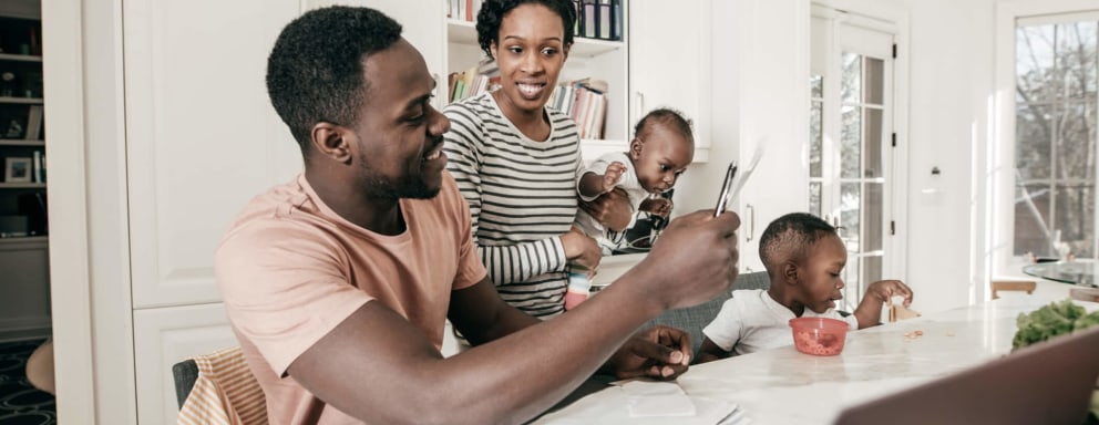 A mid-adult Black couple is going over financial documents. They are sitting at the dining table in their home with their young children.