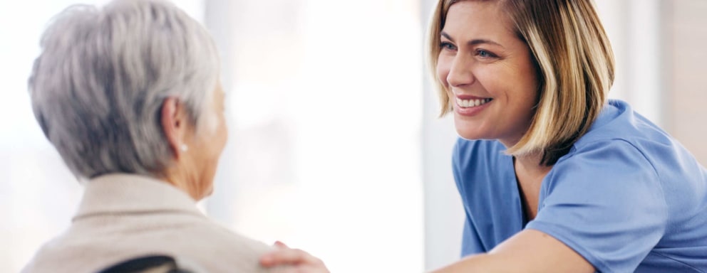 Female Caucasian nurse smiles while chatting with a senior patient in a wheelchair at a hospital.