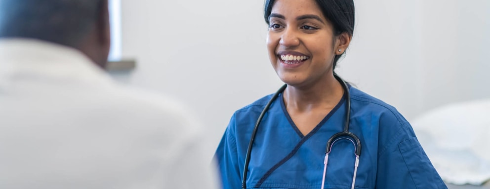 A female Indian-American nurse takes notes on a clipboard while chatting with a senior patient during an appointment. They are sitting across from each other in a clinical examination room.