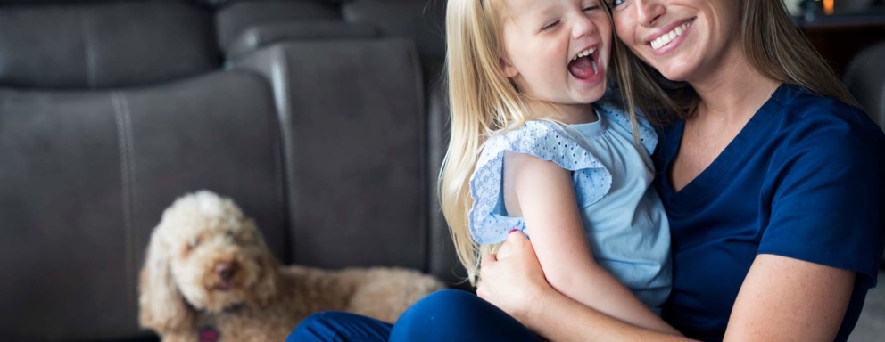 A mid-adult Caucasian nurse wearing dark blue scrubs is enjoying some bonding time with her little daughter before work. She is sitting down ion the floor of their living room, holding her daughter in her arms and smiling. The young daughter is wearing a light blue top and laughing with her eyes closed