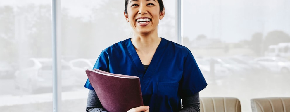 Picture of a smiling Asian-American nurse standing in a hospital waiting room. She has black hair pulled back into a ponytail, and is wearing dark blue scrubs. She is carrying a patient file in her right arm.