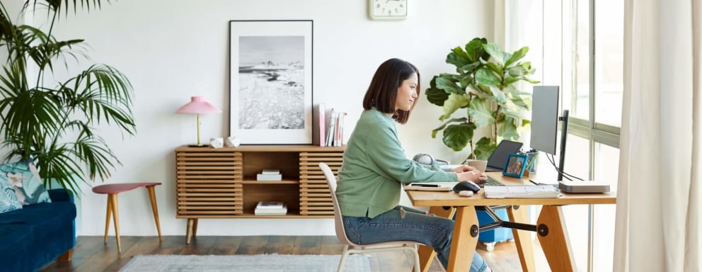 A mid-adult Hispanic woman typing on her computer at home. She is dressed casually in a sweater and jeans, and is sitting at a table in her home office.