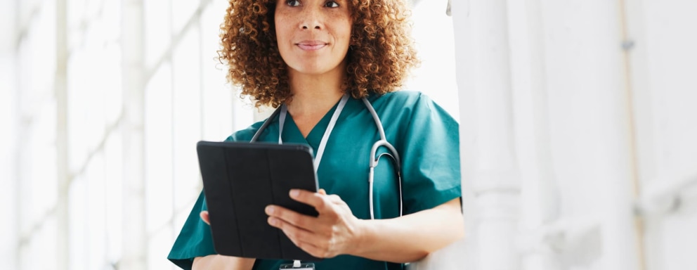 Black female nurse practitioner stands in a hospital hallway looking at patient's report on a digital tablet.