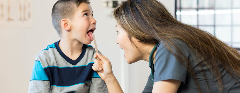 A female Asian-American school nurse examining a young boy in her office. The nurse wears dark blue scrubs and uses a tongue depressor to examine the boy's throat.