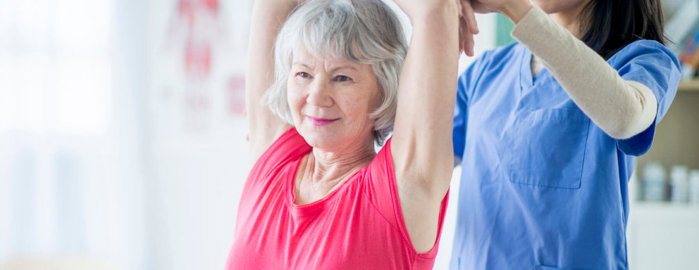 A mid-adult female holistic nurse meets with a senior adult female patient in an office at a small private clinic. The nurse is guiding the patient through some daily stretches she can do to improve her flexibility.