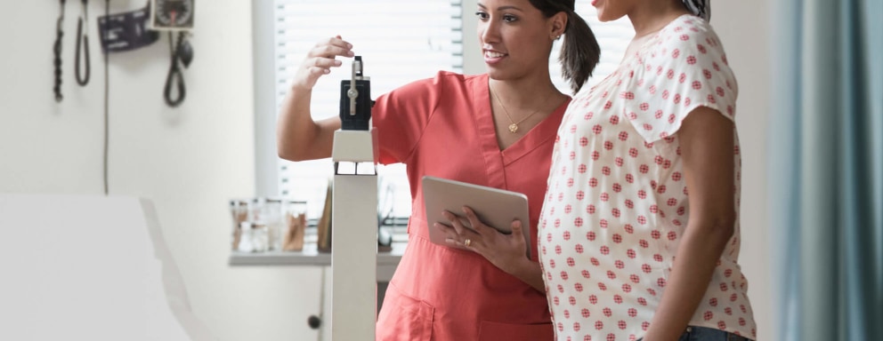 A Black female labor and delivery nurse weighs a Black pregnant patient in a hospital room.