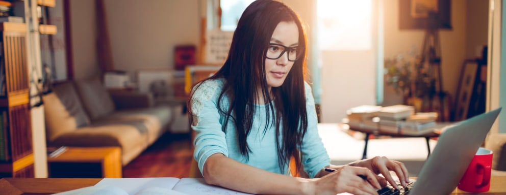 Mid-adult Asian-American woman sitting at her home desk typing on her laptop. She is wearing glasses and has a few textbooks open next to her.