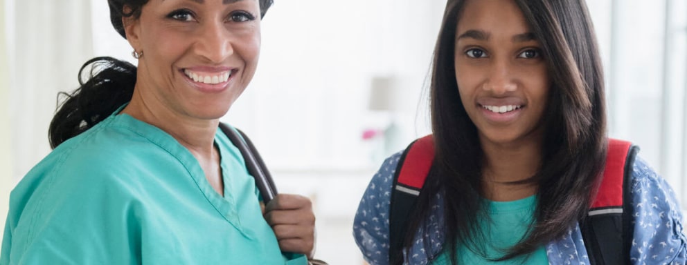 A mid-adult Black female nurse and her daughter are smiling and facing towards the camera while getting ready to leave their house in the morning. The nurse is wearing teal green scrubs and carrying a black shoulder bag over her left shoulder and a thermos of coffee in her right hand. Her teenaged daughter is wearing a light blue short-sleeved shirt and has long black hair down around her shoulders. She is wearing a black backpack with red accent trim and carrying a textbook in her hands.