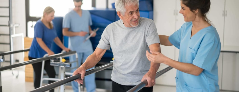 Female Hispanic nurse assisting her senior male patient with walking on the parallel bars during a therapy session. Another patient and professional are walking in the background.