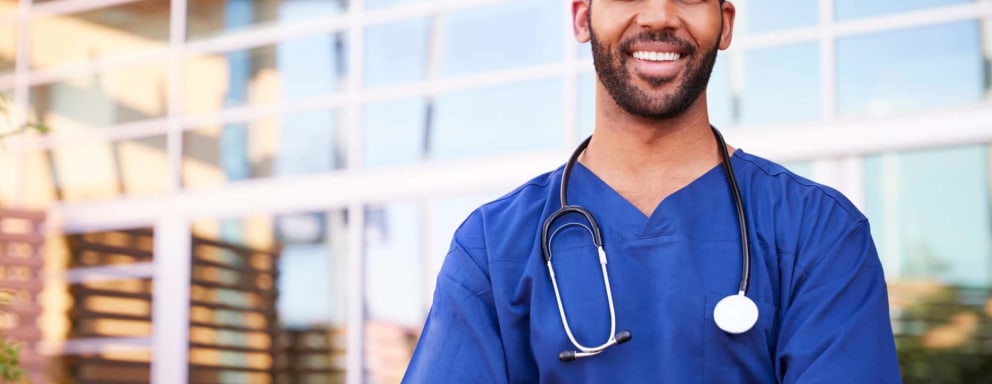 A young black male nurse standing outside a hospital smiling at the camera.