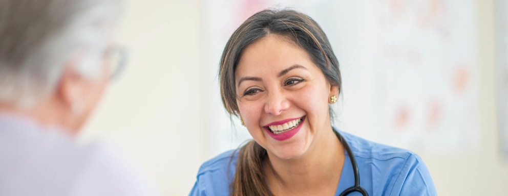 A smiling female nurse practitioner is sitting down at a table across from her senior female patient in a medical office room. The nurse is discussing the woman's current daily routine and making a revised plan of care.