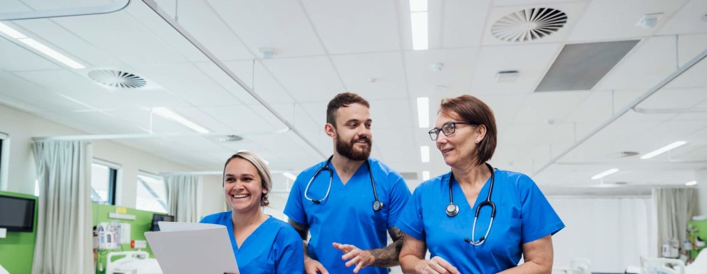 Three nurses, two female and one male, are walking and talking together through a hospital ward.