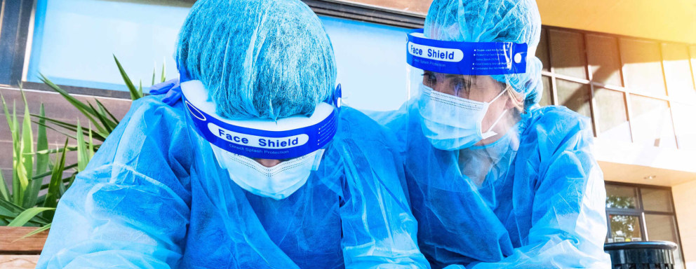 Two female nurses wearing personal protective clothing and masks sit on a bench outside of a hospital during the COVID-19 epidemic. One of the nurses is comforting her exhausted coworker by placing an arm around her shoulder.