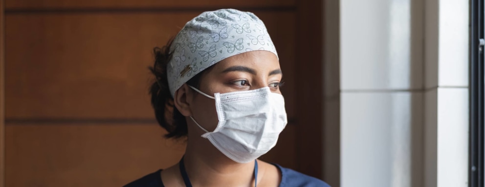 Female nurse in medical scrubs and a protective face mask taking a break from work. She is looking out from a hospital window with a tired expression on her face.