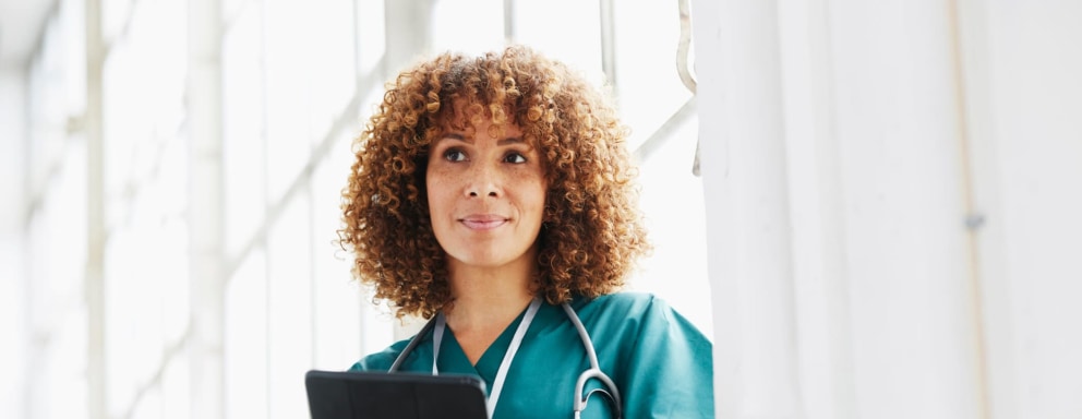 Black female nurse practitioner stands in a hospital hallway looking at patient's report on a digital tablet.