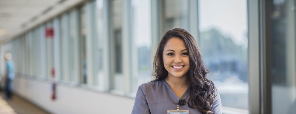 A young Filipino American nurse is standing with her arms crossed in a hospital hallway. She is wearing gray scrubs and her staff badge, and has her long black hair down around her shoulders. She is looking at the camera with a confident smile.