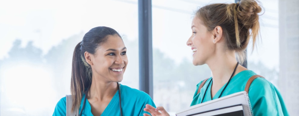 A pair of female nursing students are cheerfully chatting while walking to class together. The student pictured on the left is a young mixed-race woman with straight dark brown hair pulled back into a ponytail. She is wearing bright blue scrubs, a gray backpack, and a stethoscope around her neck, and is carrying a gray laptop in her left arm. Her classmate pictured on the right in the photo is a young Caucasian woman. She is wearing teal green scrubs and a brown backpack. She is carrying a notepad and a textbook in her left arm and a pen in her left hand. Together they are walking in a hallway lined with floor-to-ceiling glass windows with a view overlooking the school campus.