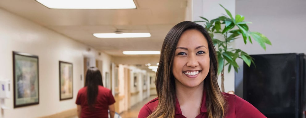 Asian-American female medical assistant smiles while standing in front of a hospital reception desk.