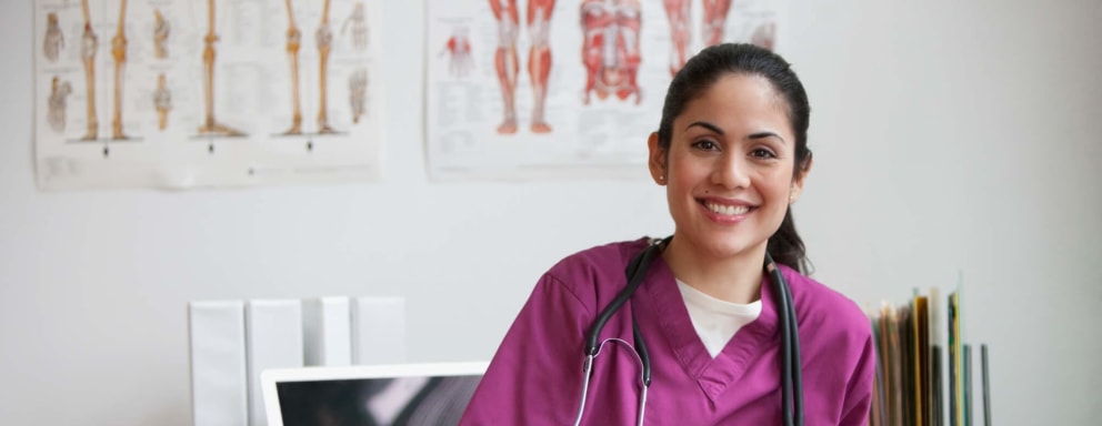 A female Hispanic nurse sitting at her desk in a clinical office. She is facing towards the camera and smiling. Her laptop and case files are on the desk behind her.