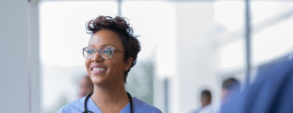 Female nurse smiles while staring out window in hospital hallway and holding digital tablet with electronic patient file