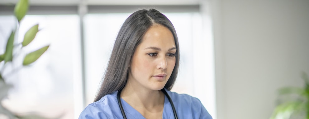 A nurse wearing scrubs interacts with a patient through a video call on her computer.