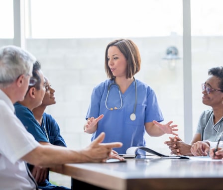 A multi-ethnic group of medical professionals are sitting together at a table discussing a public health case. A young white female nurse is leading the conversation. She has short brown hair and is wearing her light blue scrubs.