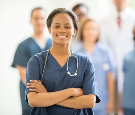 A young Black female nurse is smiling and looking at the camera. She has long brown hair pulled back into a ponytail, and is wearing dark bluish-gray scrubs. Behind her, a multi-ethnic group of doctors and nurses are standing together inside of the hospital.