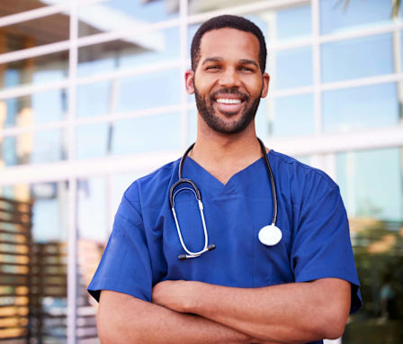 A young black male nurse standing outside a hospital smiling at the camera.