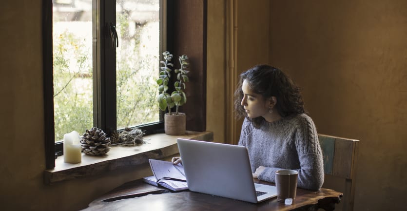 A woman sits at a dining table next to a window and consults her notebook and laptop