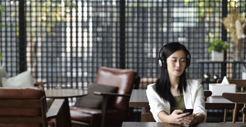 A woman with her eyes closed and headphones on sits at a table, listening to guided meditation.