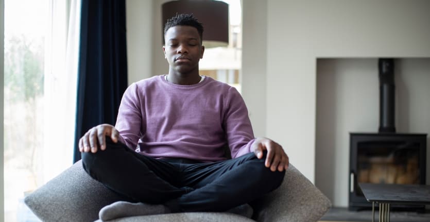 A young man sits cross-legged on a living room chair with his eyes closed in meditation.