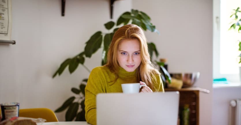 A woman in a turtleneck sips a cup of coffee as she browses the internet on her laptop.