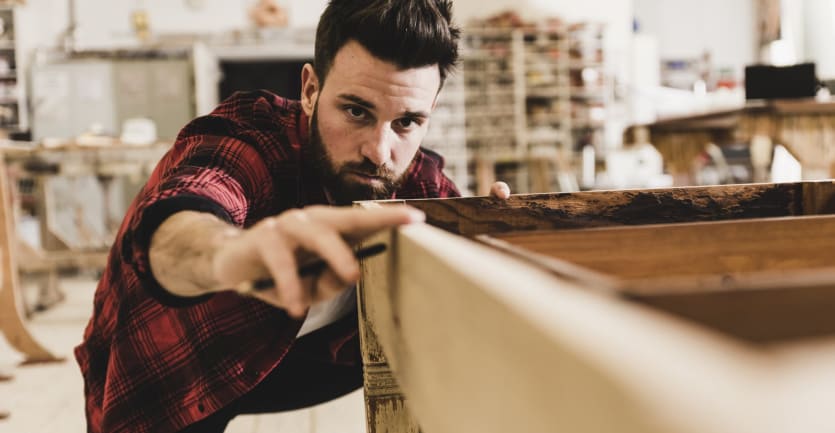 A bearded man in a workshop wearing a flannel shirt eyes a table he's restoring.