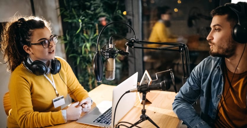 A man and woman with headphones sit at a table covered with a laptop, microphones, and other podcast equipment as they hold an earnest discussion.